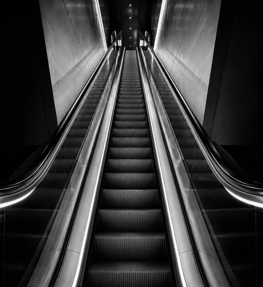 Escalator rising between lit handrails, in high-contrast black and white